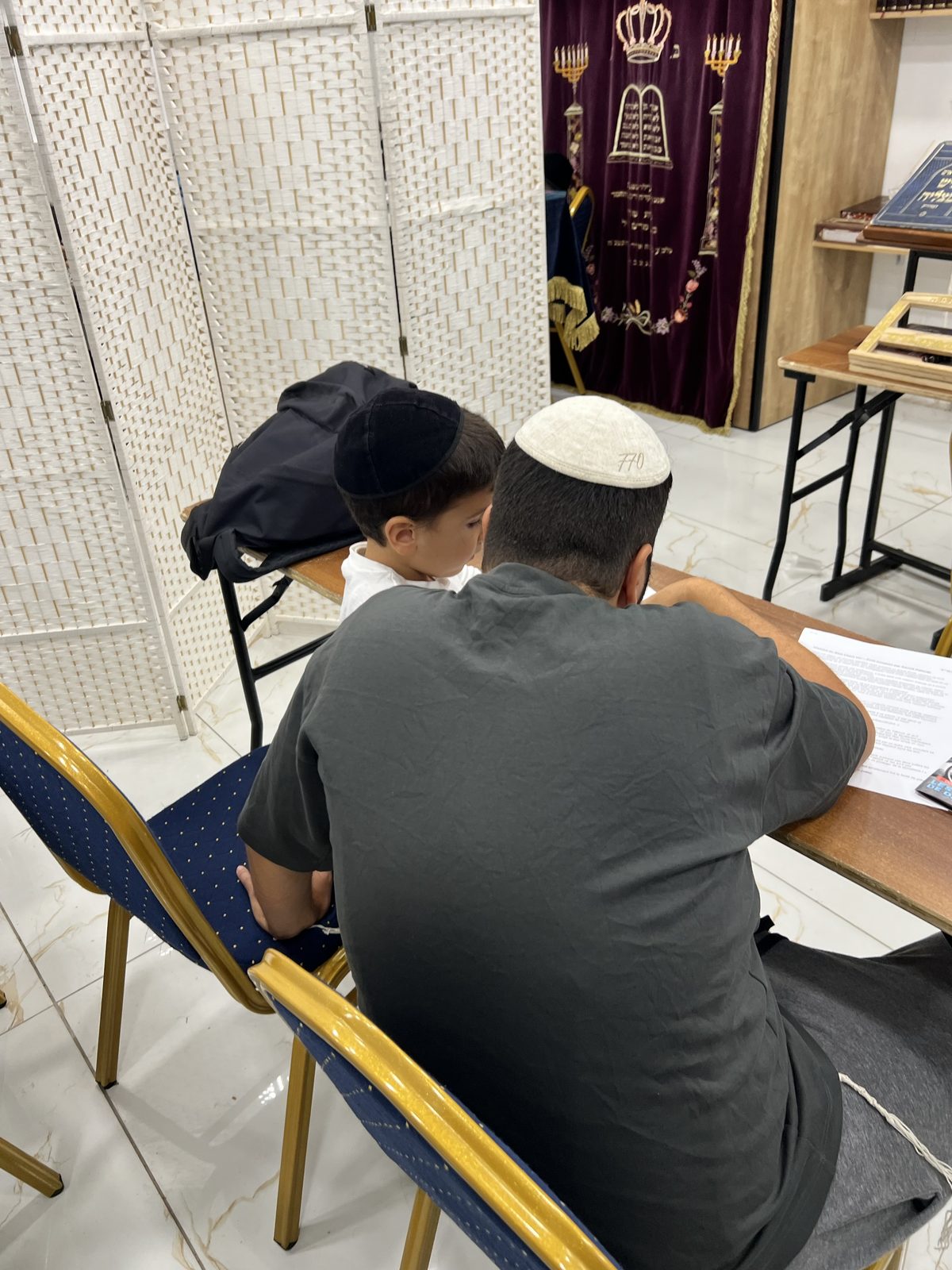 Père et fils de dos, étudiant côte à côte devant le Aron Kodesh lors d’une soirée Avot Oubanim