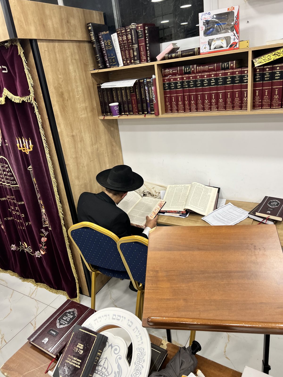 Étude au beit midrash : un homme penché sur ses sefarim, vu de dos, bibliothèque pleine