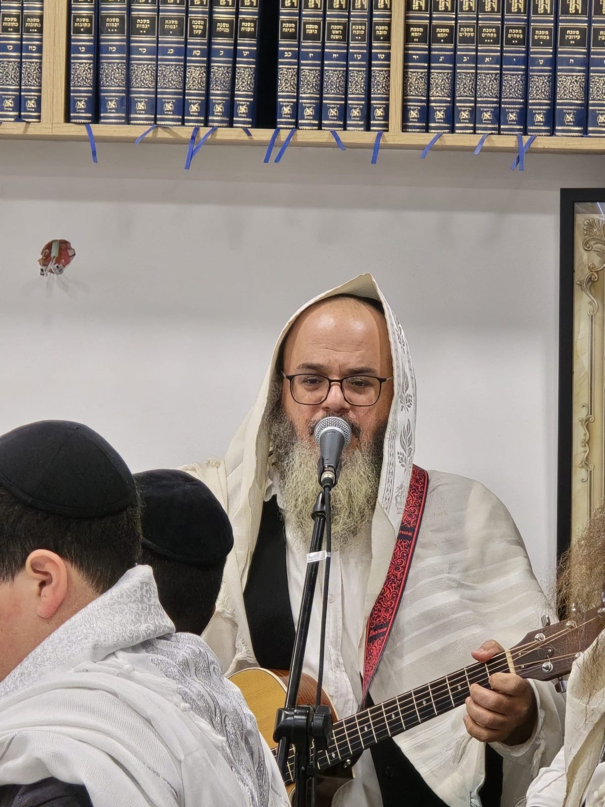 Le Rav en talit, guitare en main, chantant au micro devant la bibliothèque de sefarim